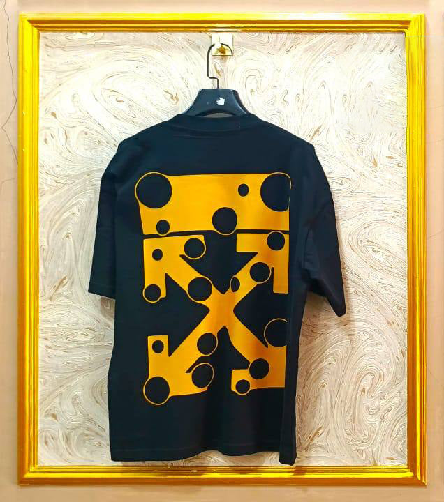 Black t-shirt with yellow geometric pattern on a hanger against a decorative wall.